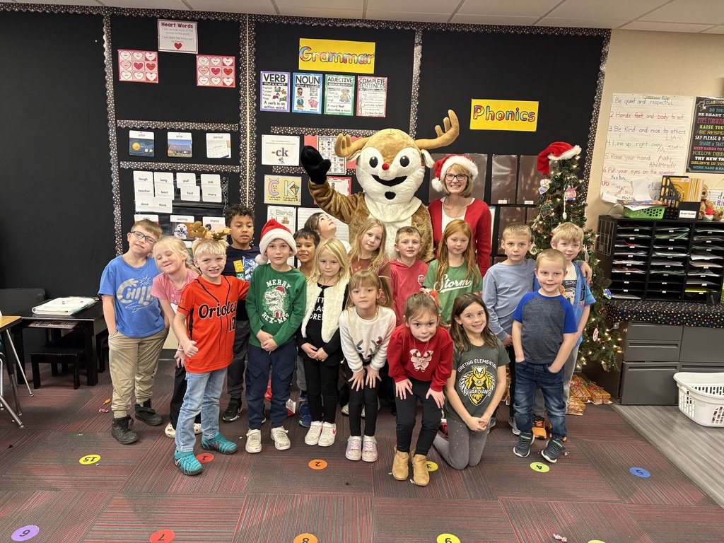 Group of students standing in a classroom around a person dressed as Rudolph and a woman with a Christmas outfit  in the front of the room next to a Christmas tree.