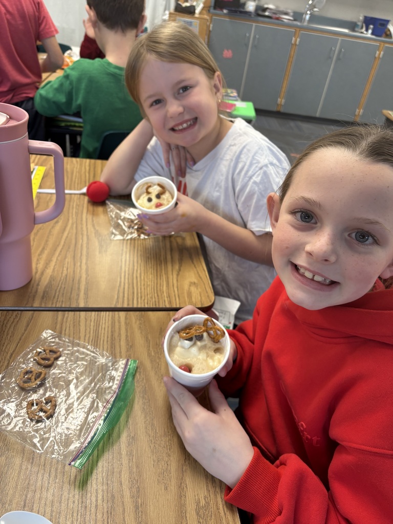 Two girls  smile at desks with a cup with a reindeer dessert inside.