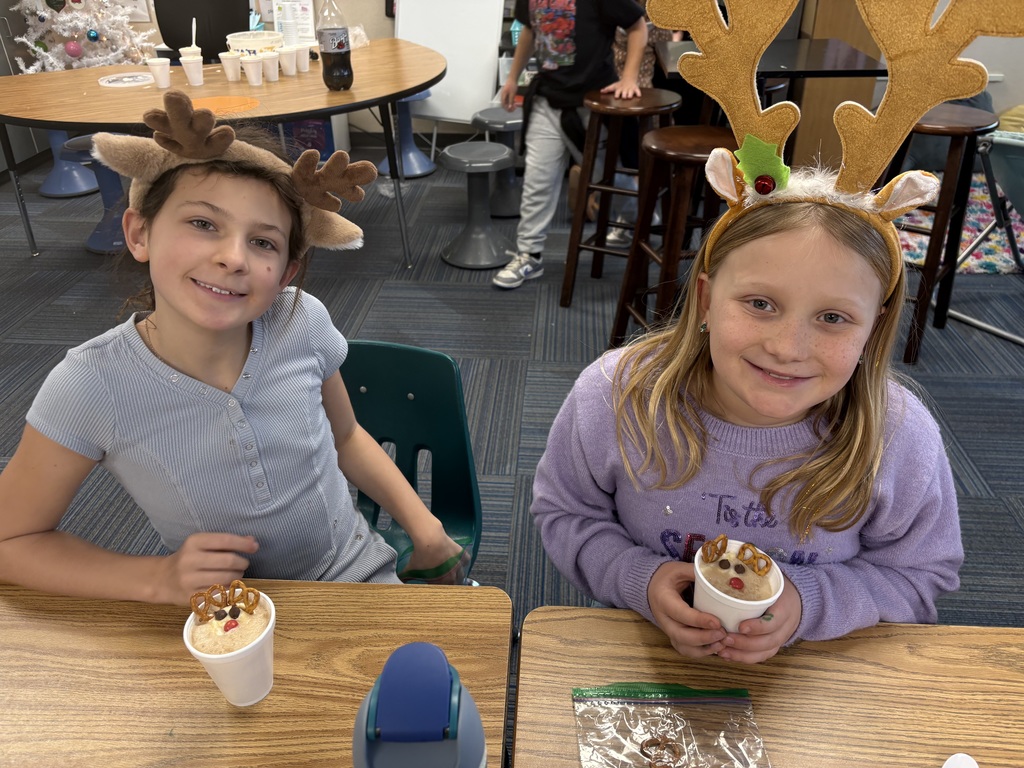 Two girls wearing reindeer hats smile at desks with a cup with a reindeer dessert inside.