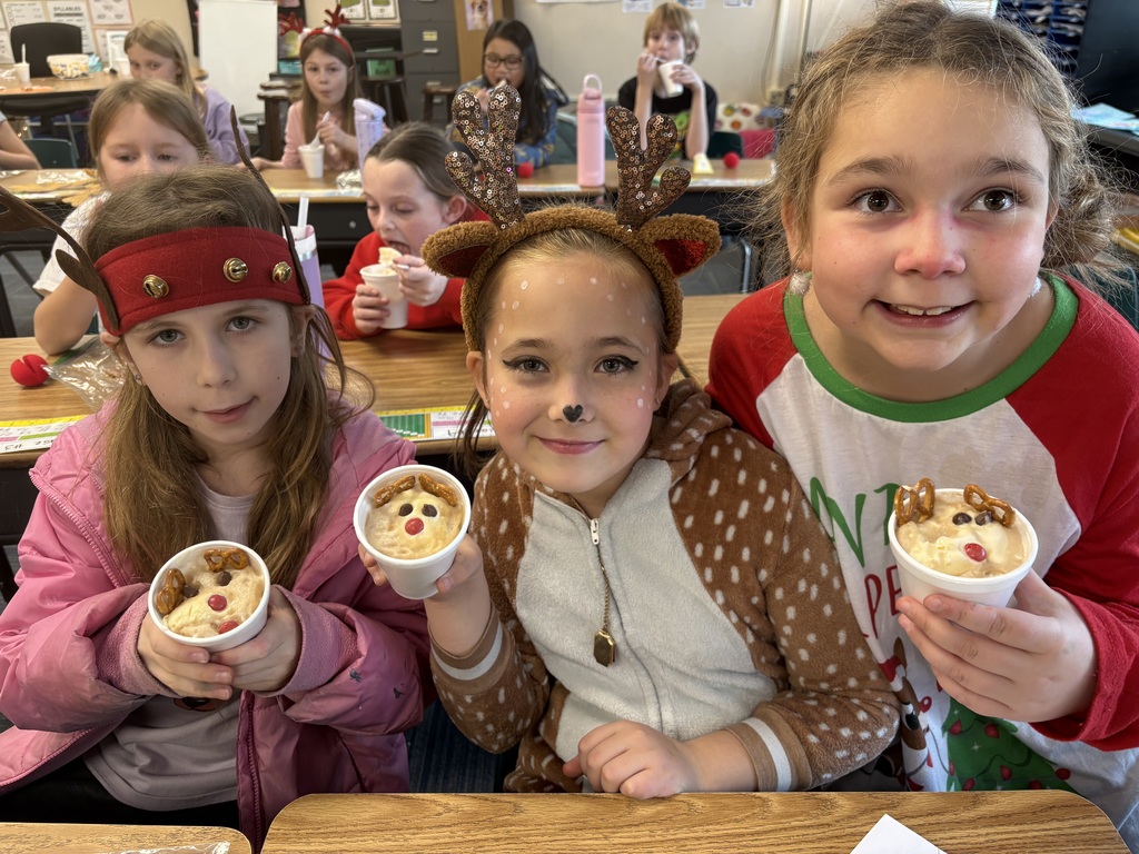 Three girls wearing reindeer hats smile at desks with a cup with a reindeer dessert inside.