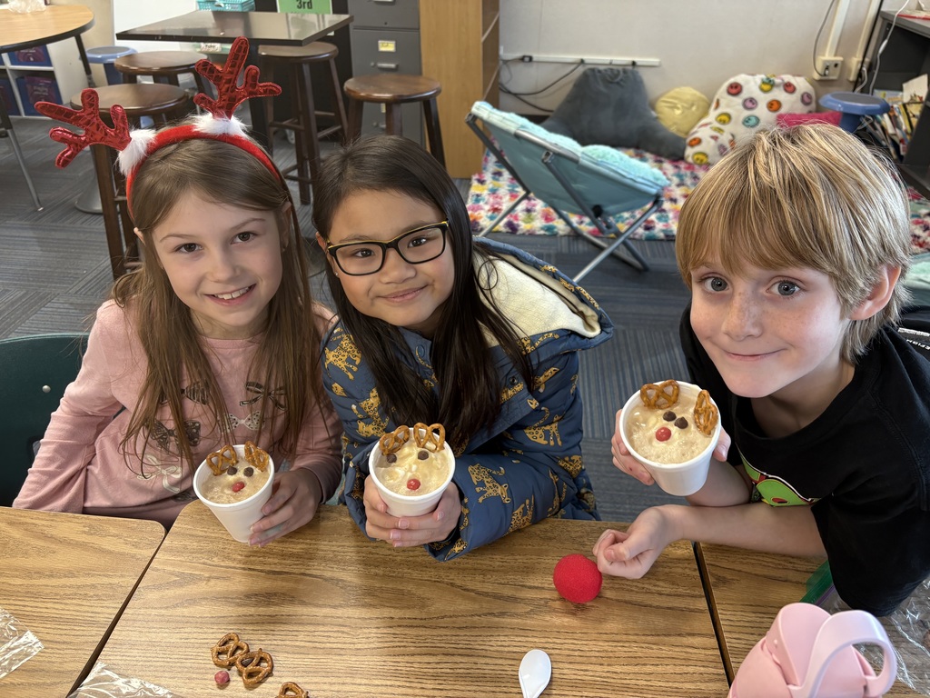 Three students wearing reindeer hats smile at desks with a cup with a reindeer dessert inside.