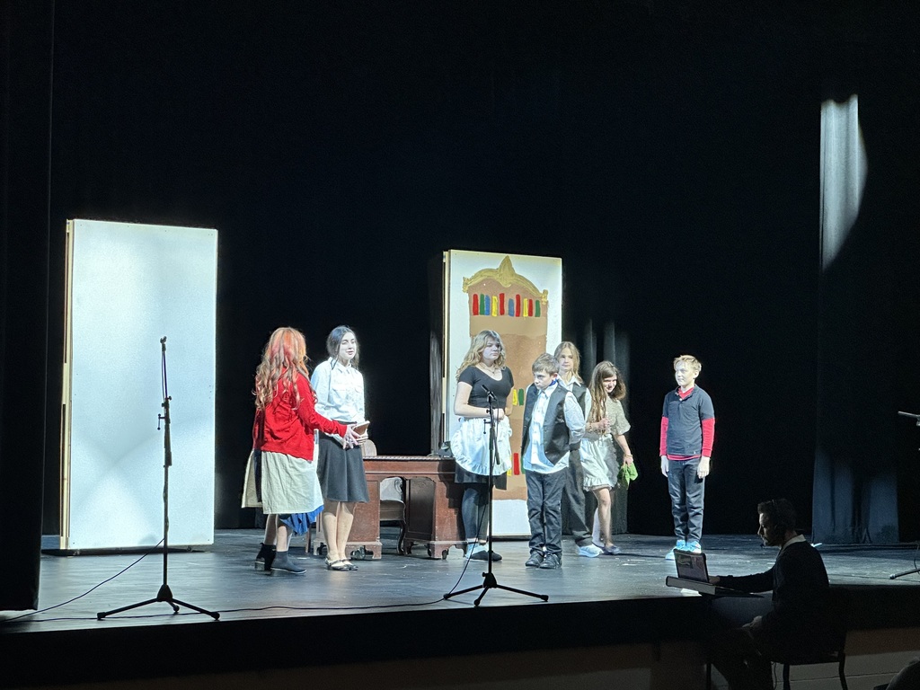 There are a group of student actors on a stage. . There is a man sitting in front of the stage playing the music.