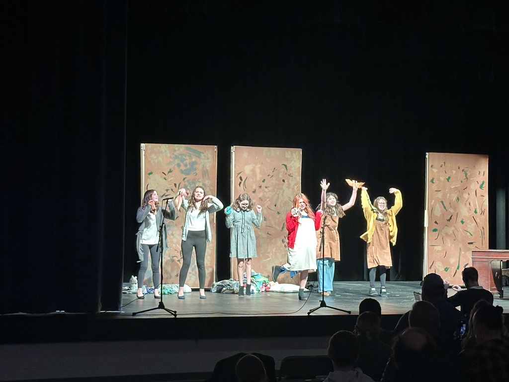 There are a group of student actors on a stage. There is a man sitting in front of the stage playing the music.