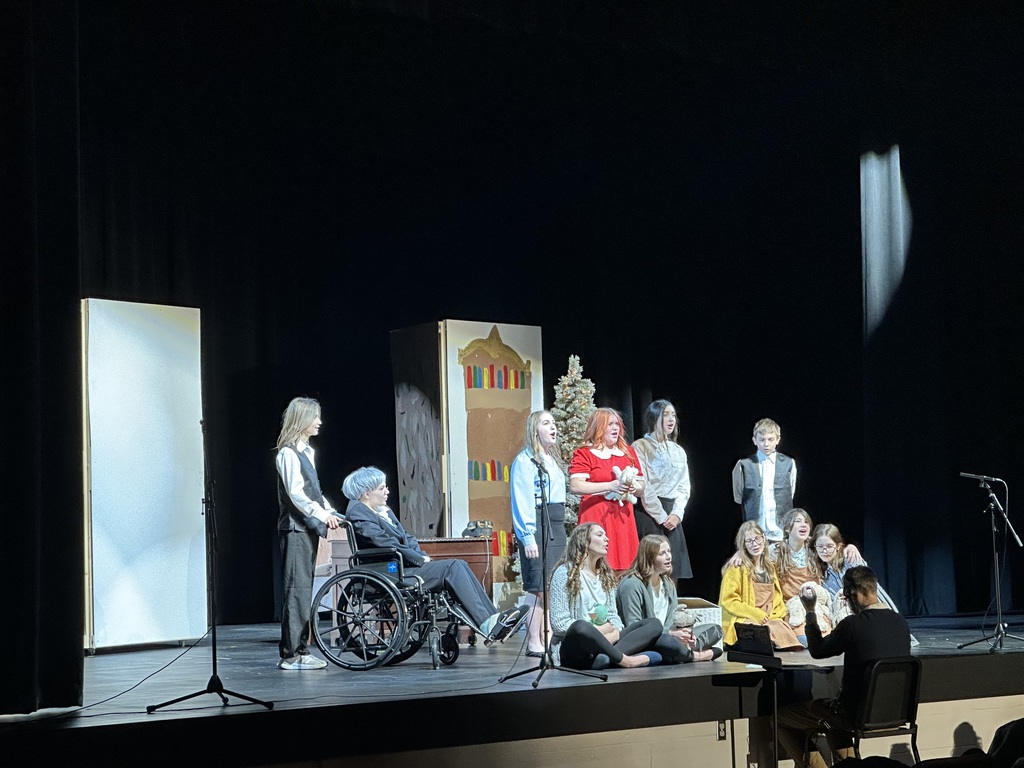 There are a group of student actors on a stage. Some are seated, some are standing, and one is in a wheelchair. There is a man sitting in front of the stage playing the music.