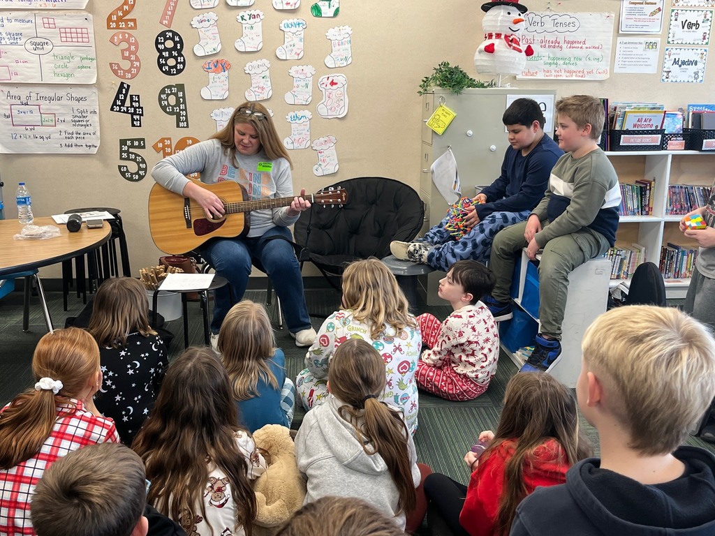 Woman sitting in the front of the room playing a guitar while students sit around the room watching her.