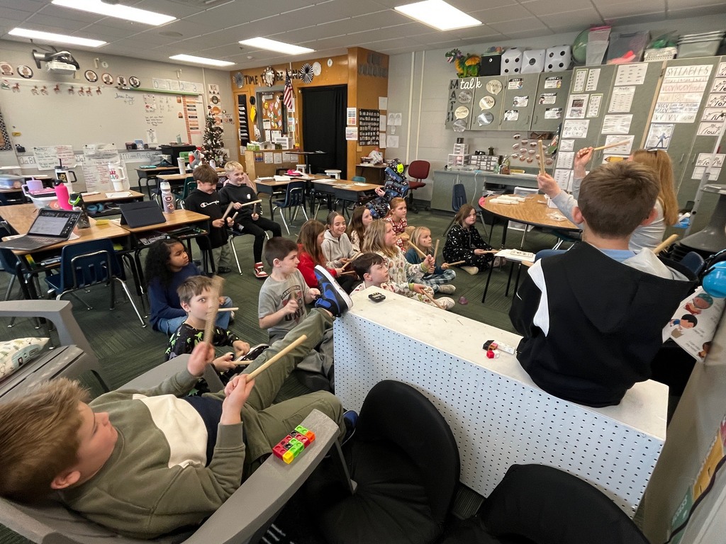 Group of students in a classroom sitting around the room holding up drumsticks while a woman presents in the front of the room.