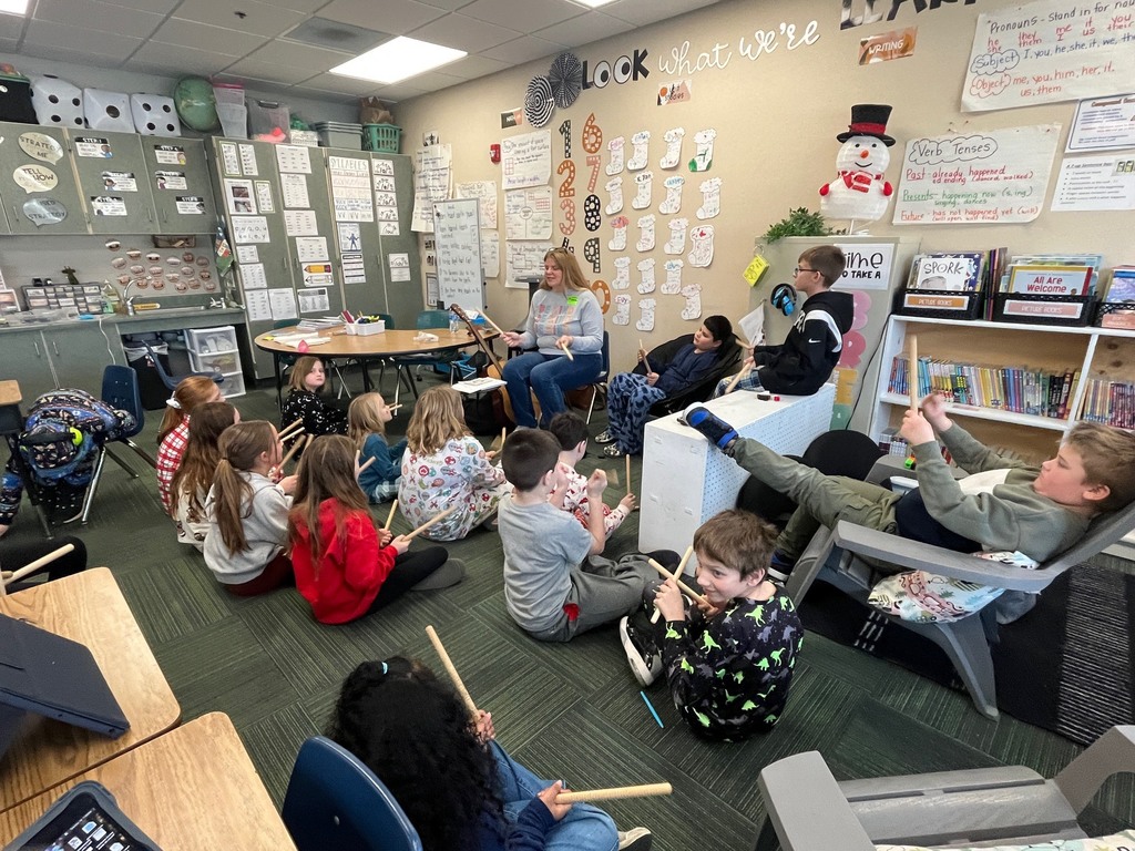 Group of students in a classroom sitting around the room holding up drumsticks while a woman presents in the front of the room.