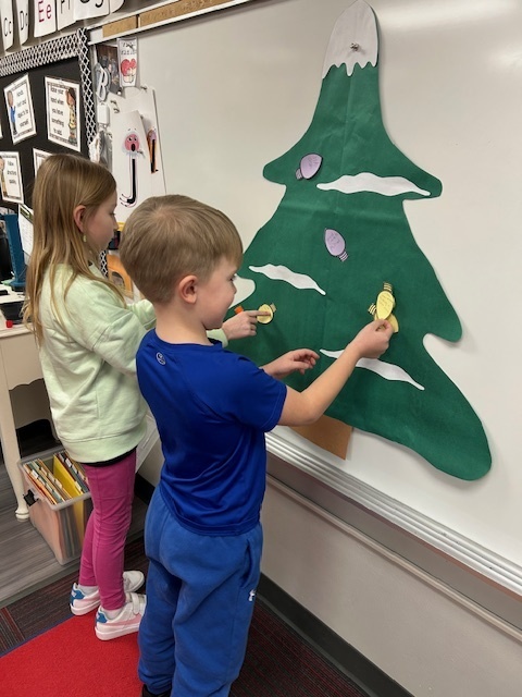 Two students stand in the front of the room putting Christmas light cutouts on a green Christmas tree.