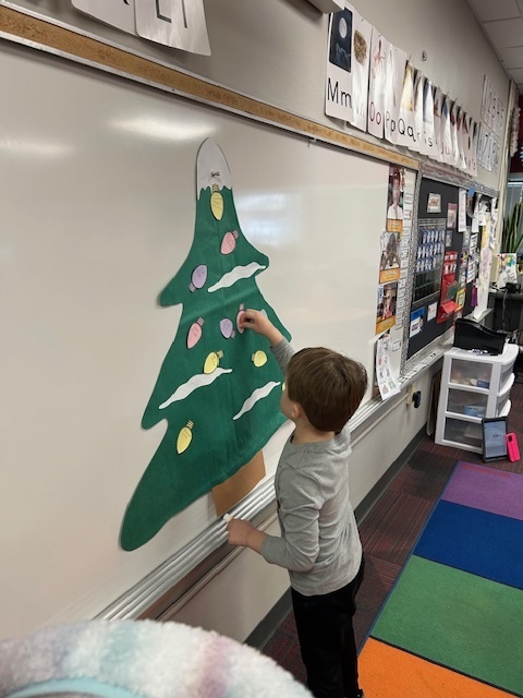 A boy standsin the front of the room putting Christmas light cutouts on a green Christmas tree.