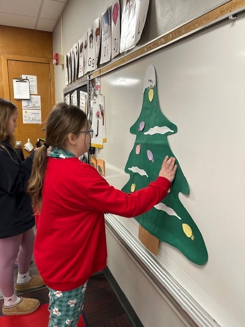 Two students stand in the front of the room putting Christmas light cutouts on a green Christmas tree.