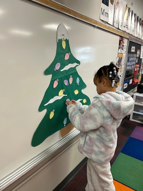 A girl stands in the front of the room putting Christmas light cutouts on a green Christmas tree.
