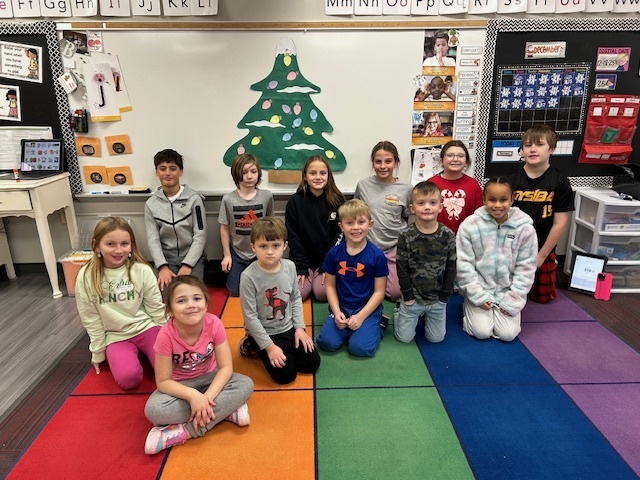 Group of students kneeling on a classroom floor with a Christmas tree cutout visible on the whiteboard behind them.