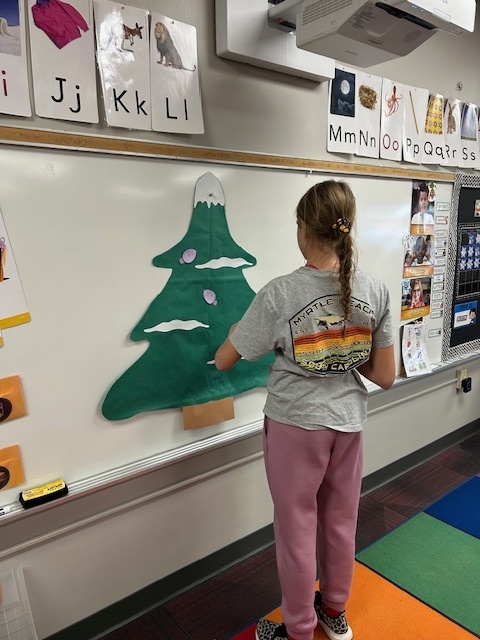 One girl stand in the front of the room putting Christmas light cutouts on a green Christmas tree.