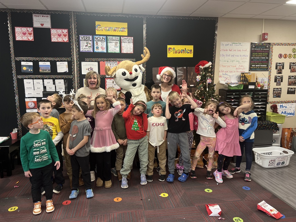 A group of students and their teacher stand in a classroom making silly faces around a person dressed in a Rudolph costume and a woman with a Santa hat.