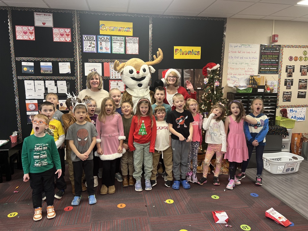 A group of students and their teacher stand in a classroom around a person dressed in a Rudolph costume and a woman with a Santa hat.