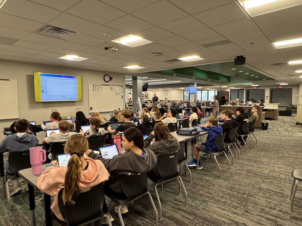 Students sit at tables looking at a TV projecting the videos. In the background another class is following along with a different TV projecting the same video.