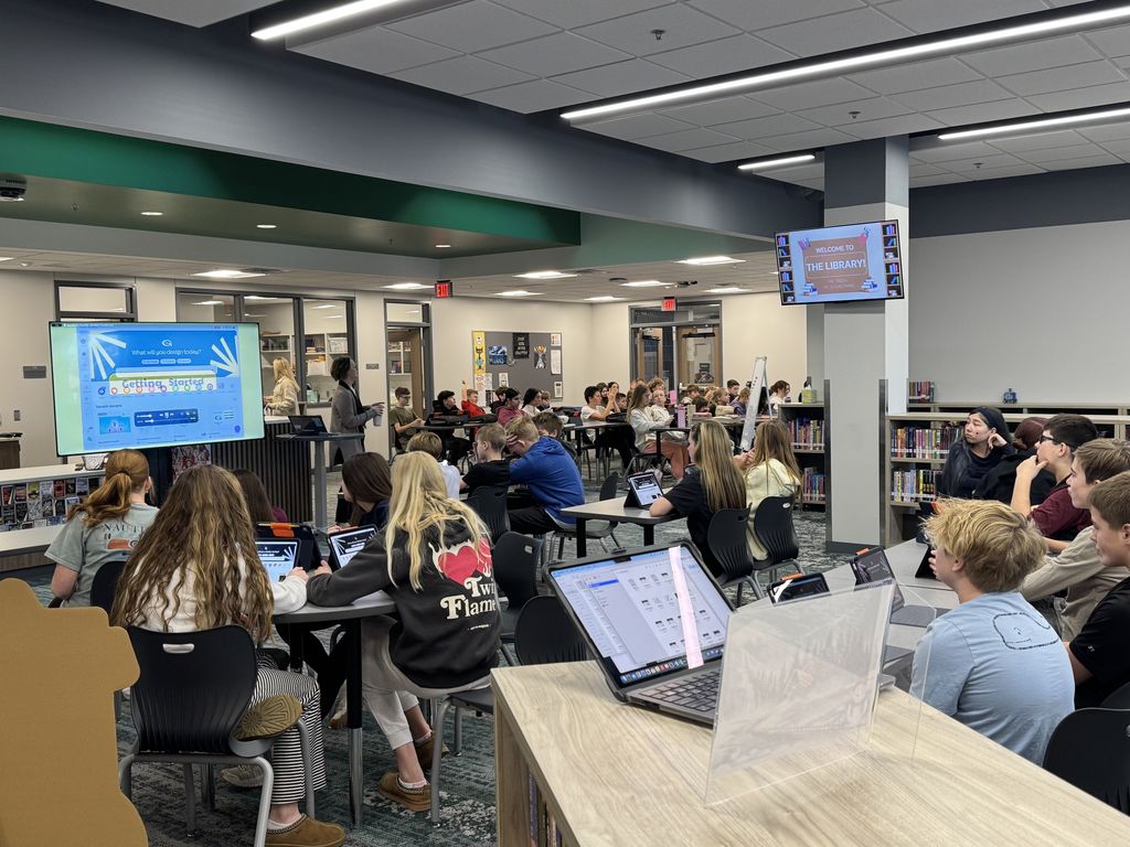 Students sit at tables looking at a TV projecting the videos. In the background another class is following along with a different TV projecting the same video.