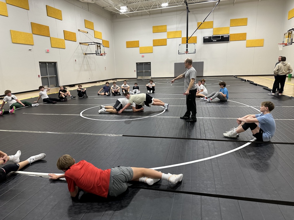 A group of boys sit in a circle on a wrestling mat, watching two male wrestlers in the center. A coach is instructing. 