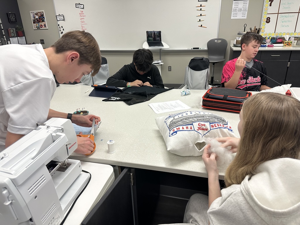Photo of four students sitting at a table working on their pillows. Two are sewing. One is cutting, and the last is stuffing the pillow.