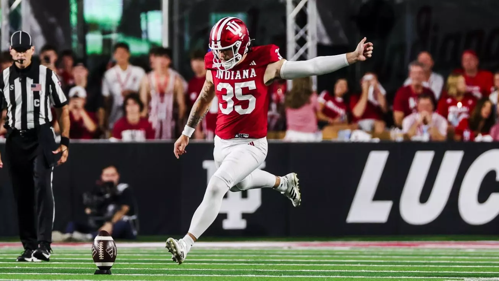 Football player wearing a red jersey and white pants kicking a football.