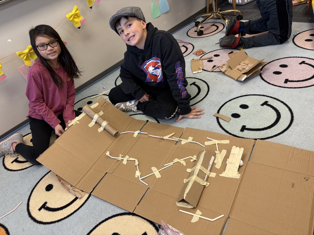 Two students smiling while kneeling on the ground in front of a cardboard engineering design