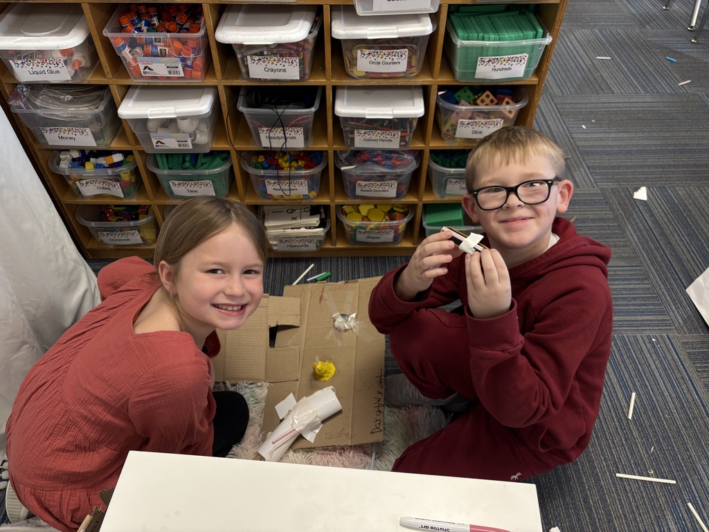 Two students smiling while kneeling on the ground in front of a cardboard engineering design
