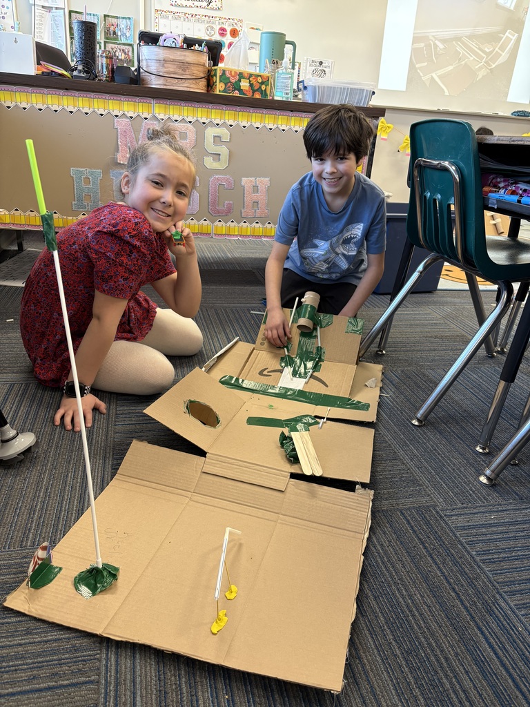 Two students smiling while kneeling on the ground in front of a cardboard engineering design