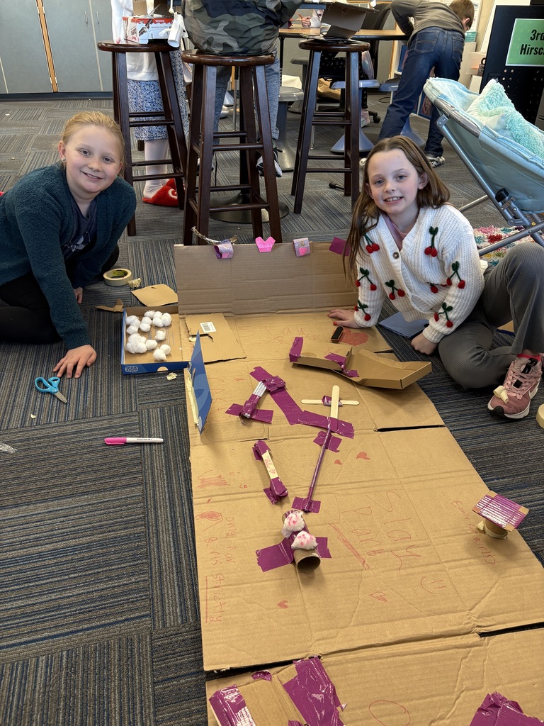 Two students smiling while kneeling on the ground in front of a cardboard engineering design