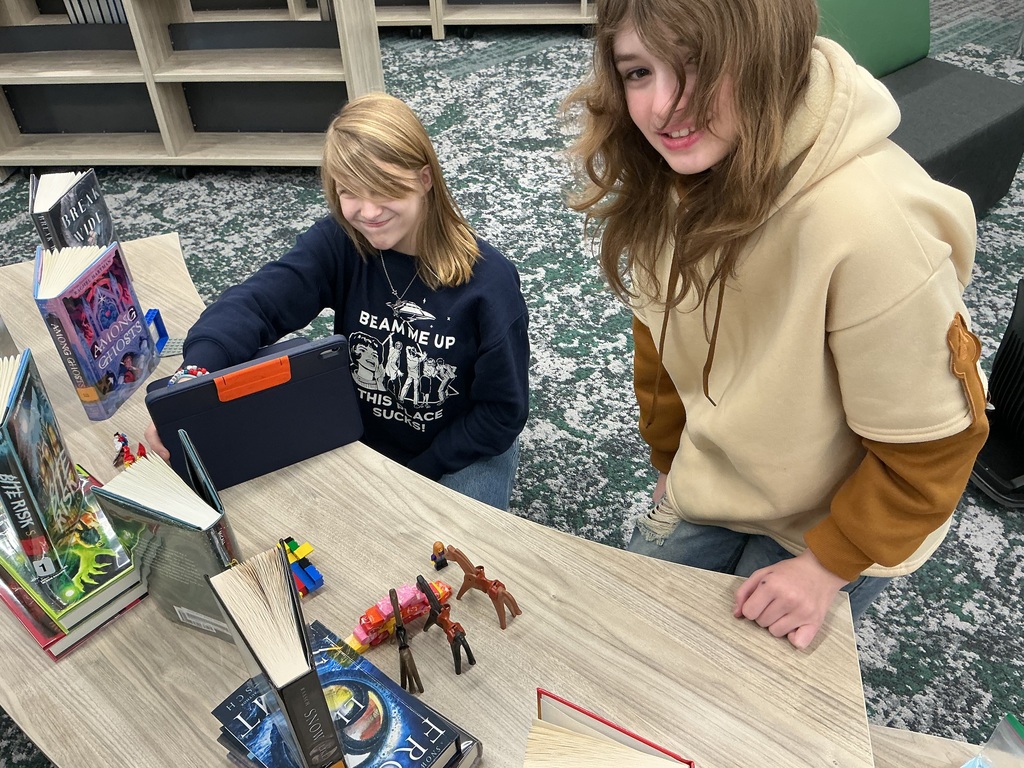 Two girls are at a table with an ipad and toys working on a project. 