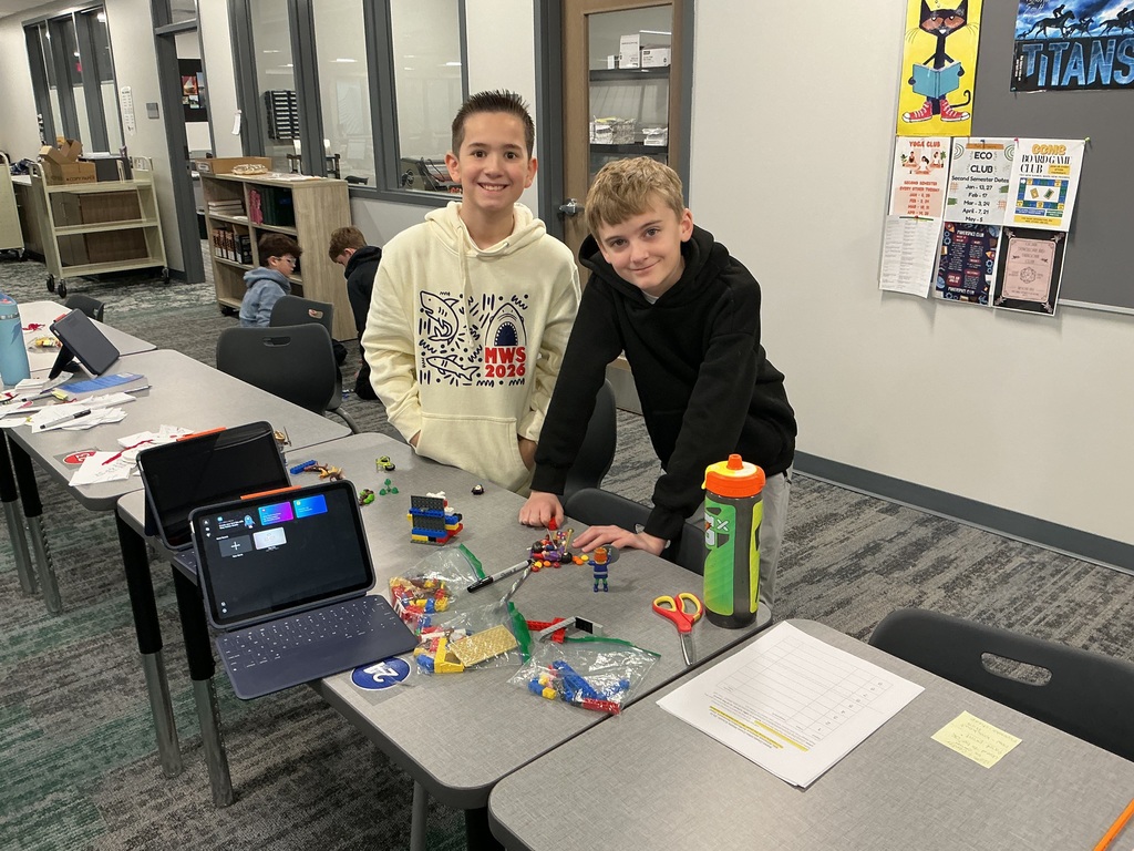 Two boys are a table with an iPad and toys working on a project. They are smiling at the camera. 