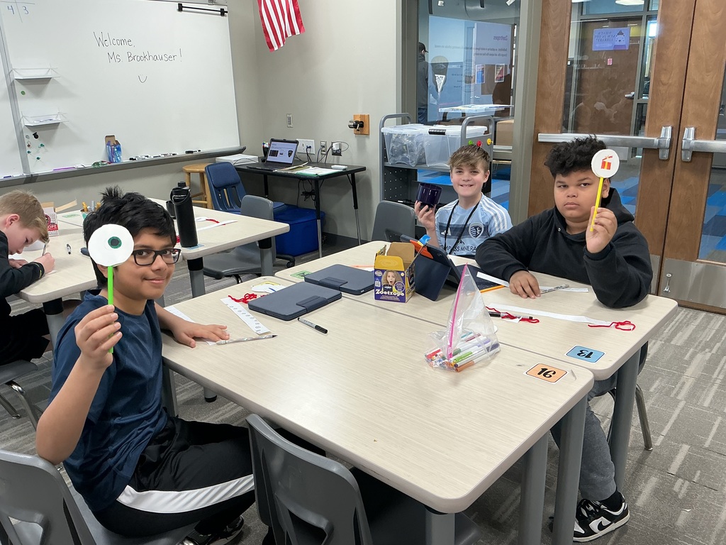 Photo of three male students sitting at a table holding up their creations of images of circles attached to the end of pipe cleaners.