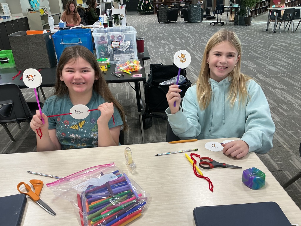 Photo of two female students sitting at a table holding up their creations of images of circles attached to the end of pipe cleaners.