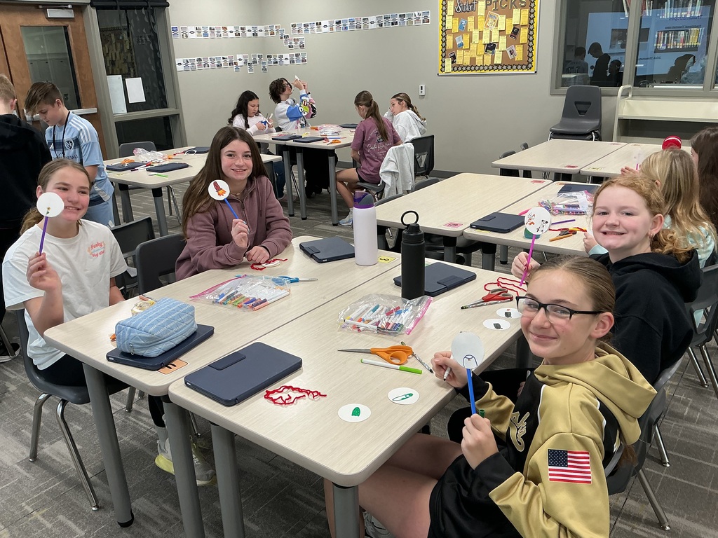 Photo of four female students sitting at a table holding up their creations of images of circles attached to the end of pipe cleaners.