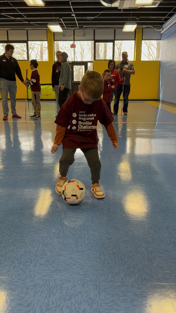 Student kicks a soccer ball on a tile floor while others watch in background.