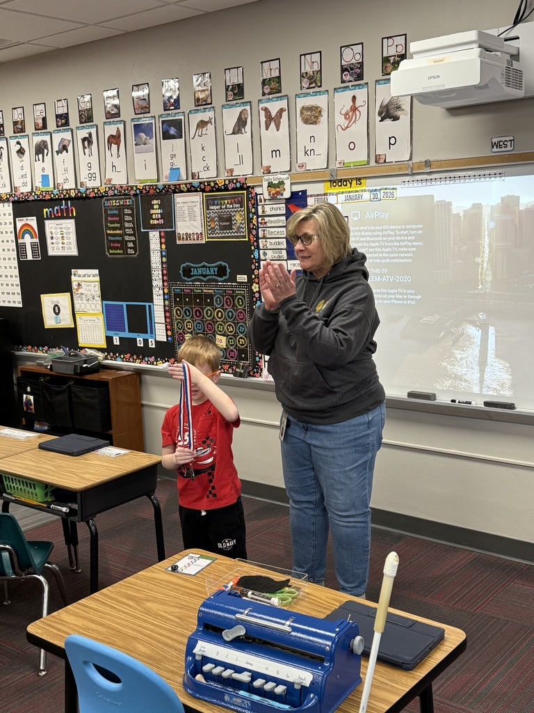 Student stands at the front of the room holding a medal while his teacher claps.