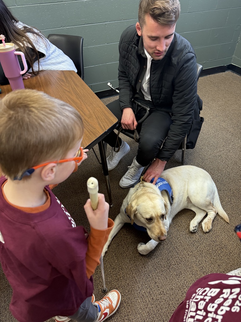 Student holds a white cane while talking to an adult who is sitting petting a service dog.
