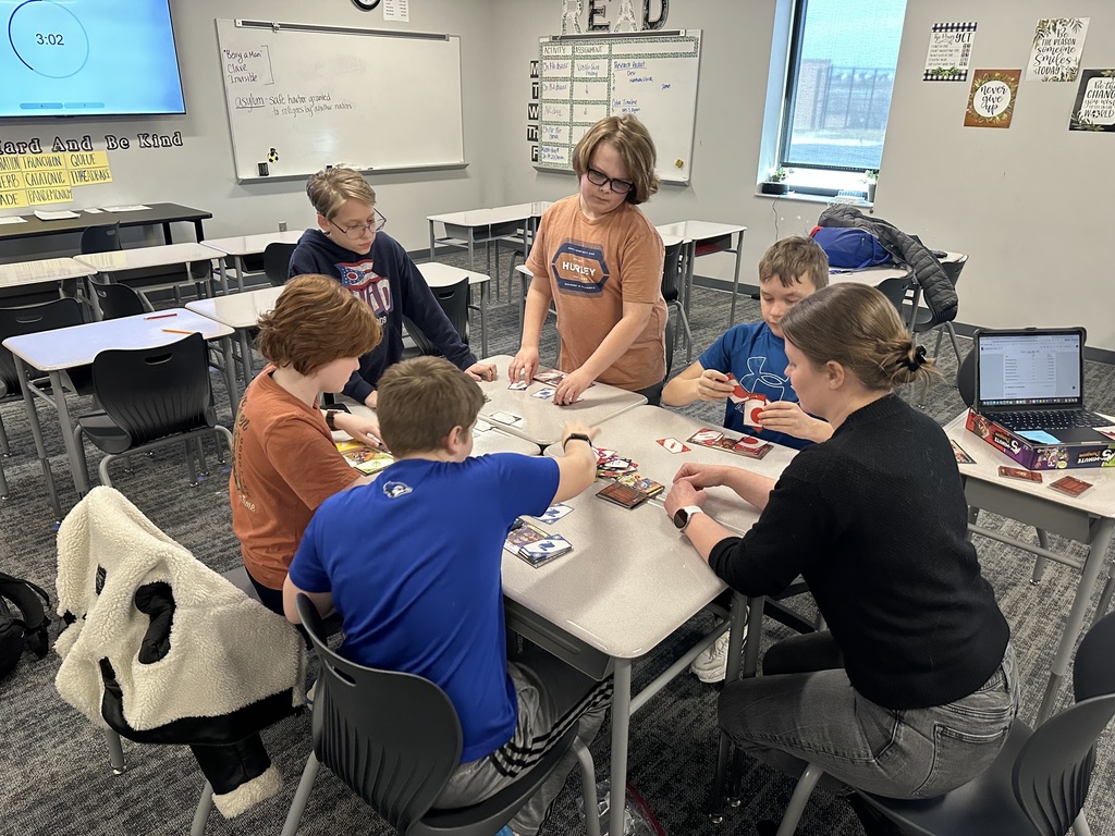 Five students and one teacher sit around a group of desks playing a board game. 