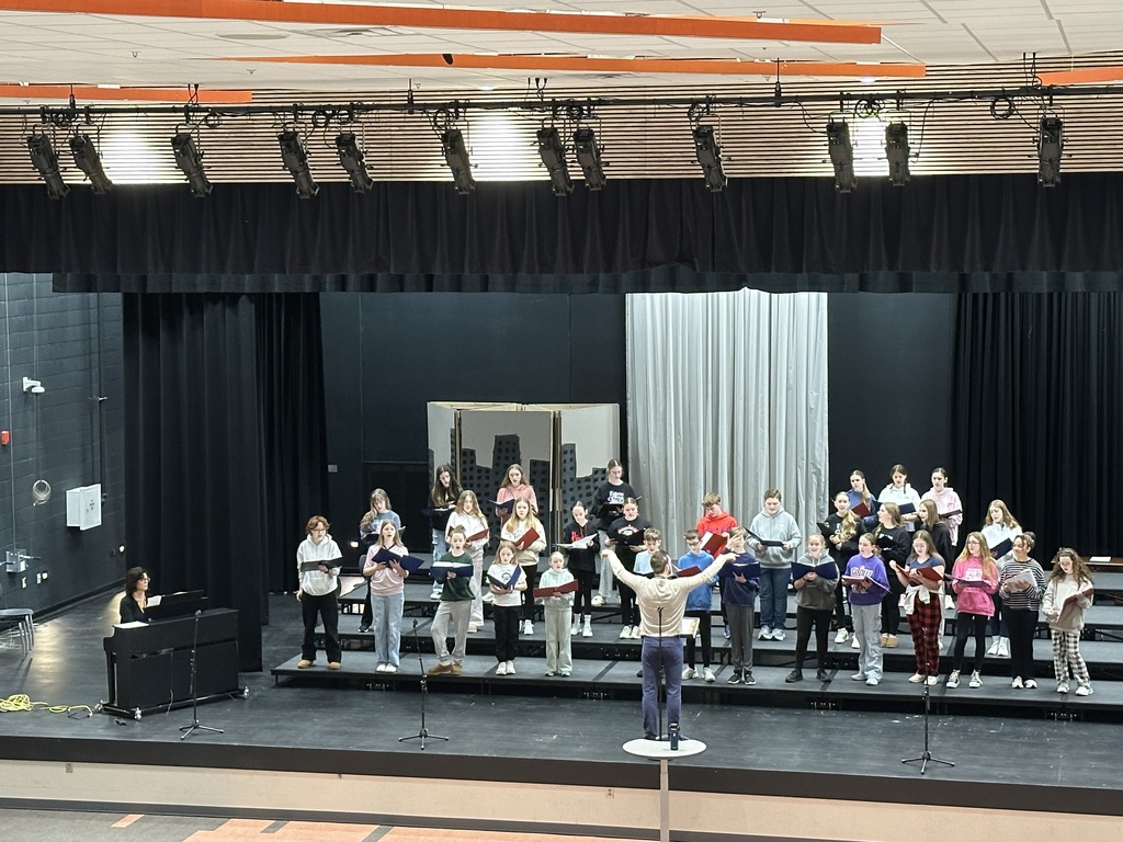 A large group of students are on risers on a stage with a male teacher directing and a woman accompanying them on the piano. 