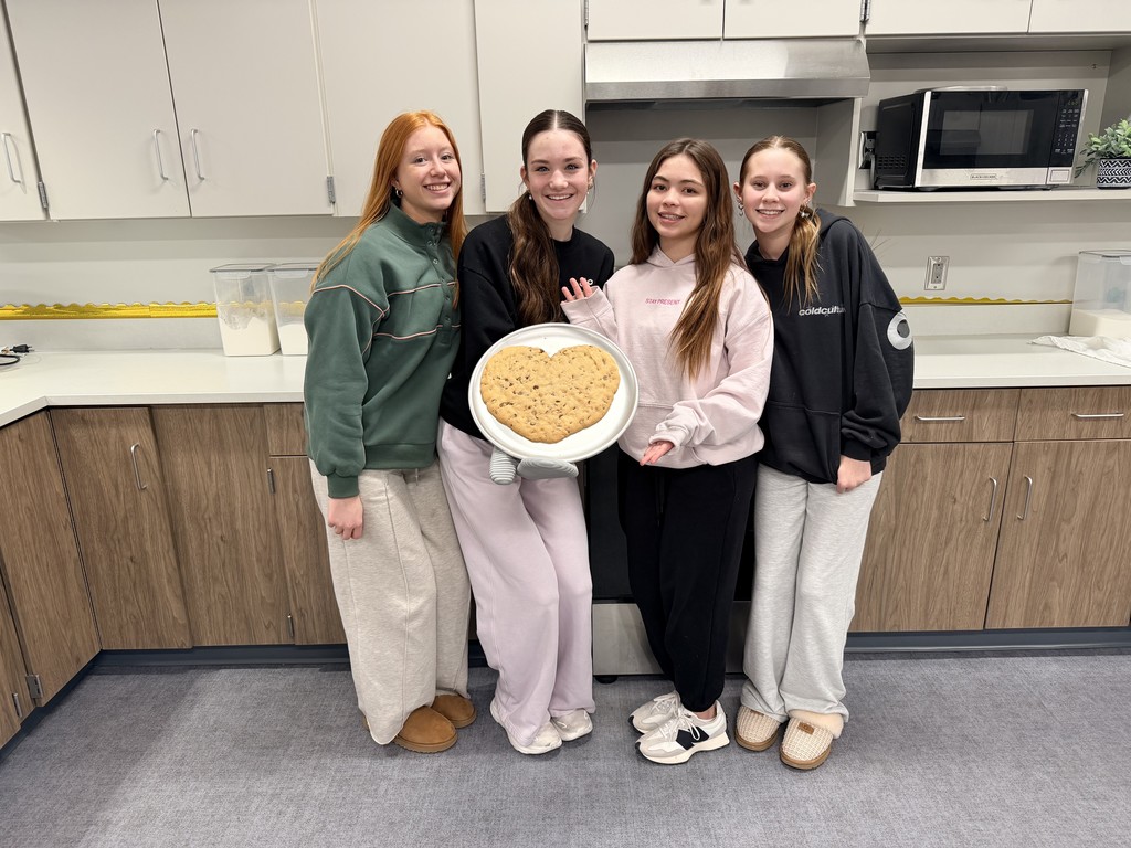 Four female students pose with a heart shaped cookie in a kitchen. 