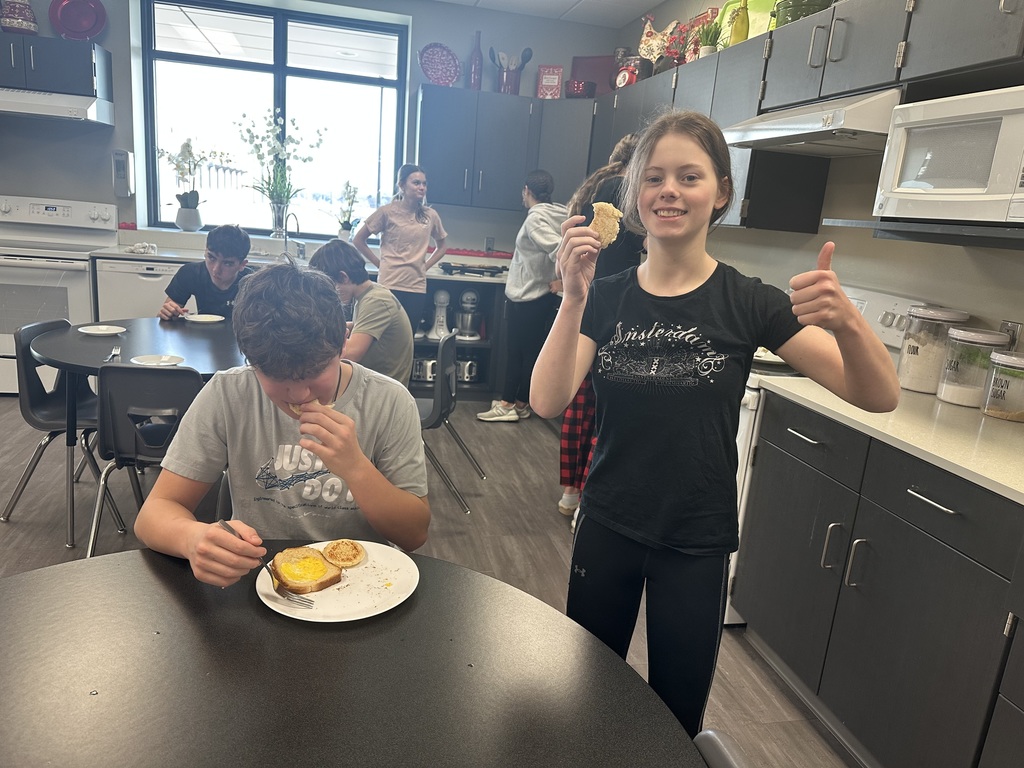Photo of a male student sitting at the table eating and a female standing next to him with toast in one hand and a thumbs up on the other.