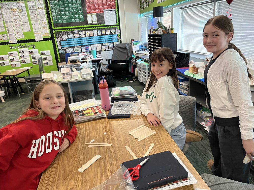 Three girls, sitting and standing around a table working on a craft with popsicle sticks.
