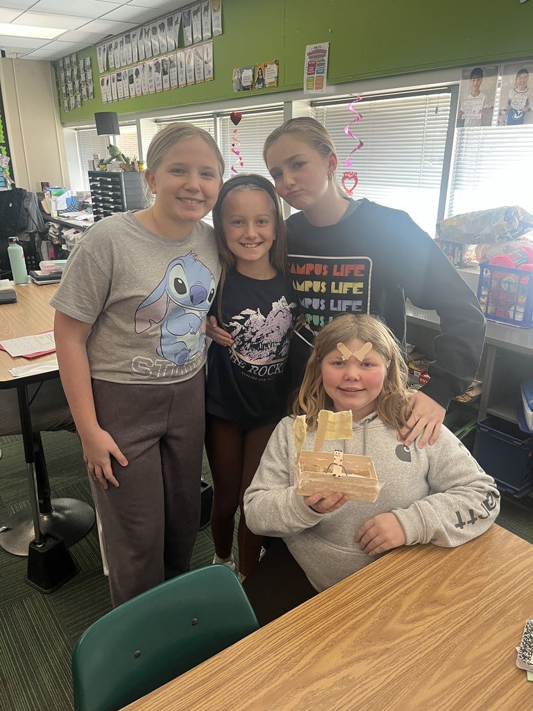 Four girls, one sitting at a table holding a craft made out of popsicle sticks and three standing, 