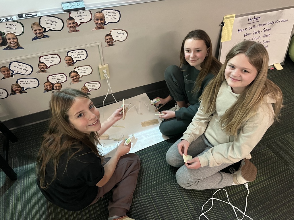 Three girls sitting on a classroom carpet working on a craft with popsicle sticks and hot glue.
