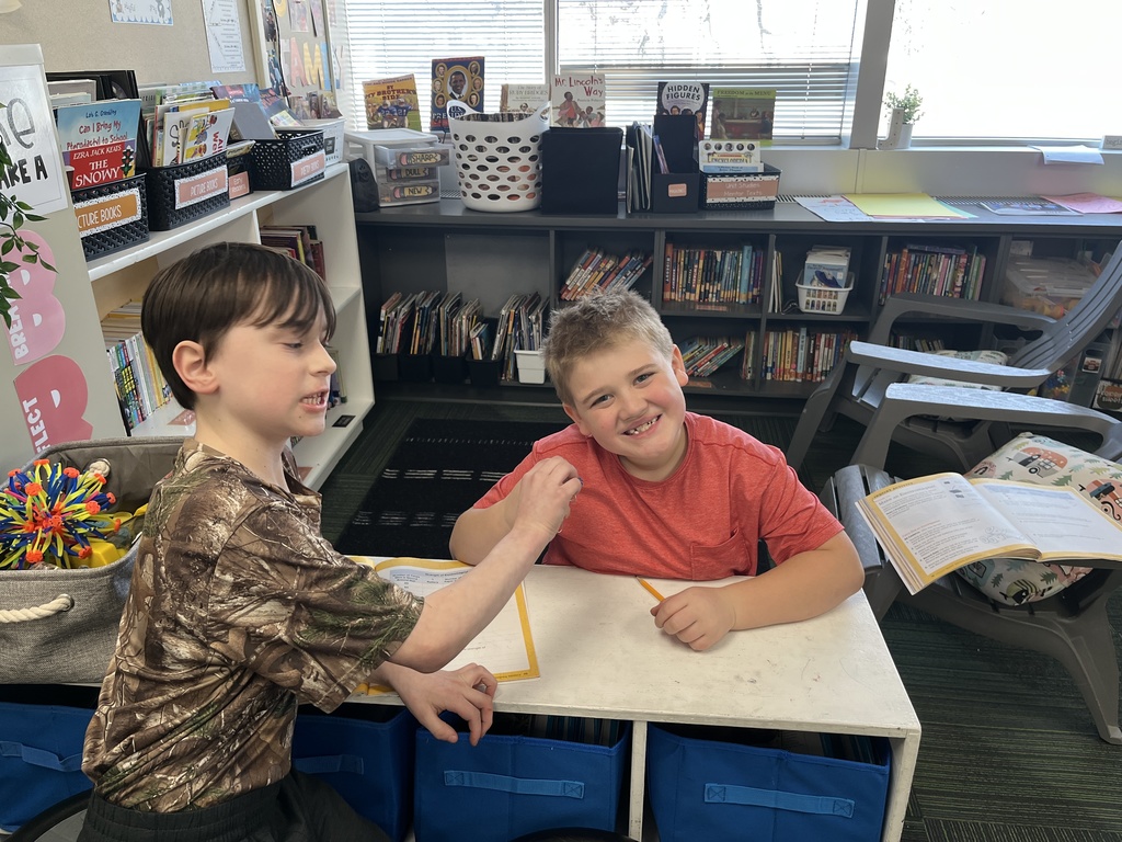 Two boys sitting at a desk smiling holding something in their hand