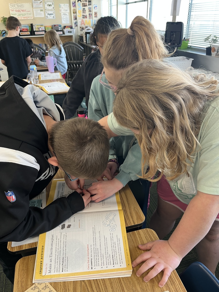 Three students stand around a desk holding paper clips while looking at a book