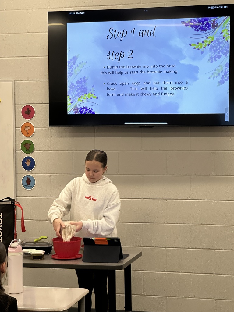 A female student is pouring brownie batter mix into a bowl in front of the class. She has a slide behind her with directions. 