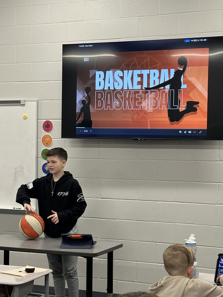 A student is showing a basketball in front of his class and has a slide behind him labeled "Basketball" .