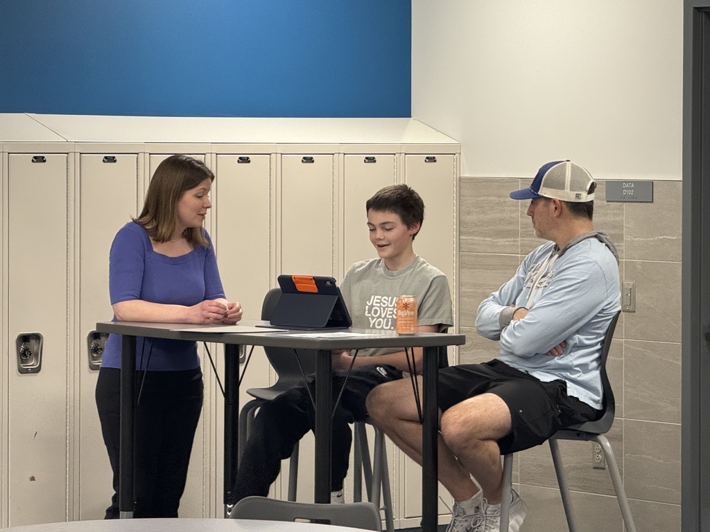 Students and parents sit at desks in a classroom chatting with teachers. 