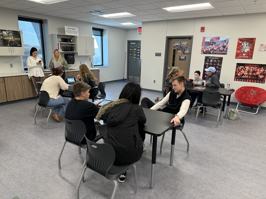 Students and parents sit at desks in a classroom chatting with teachers. 