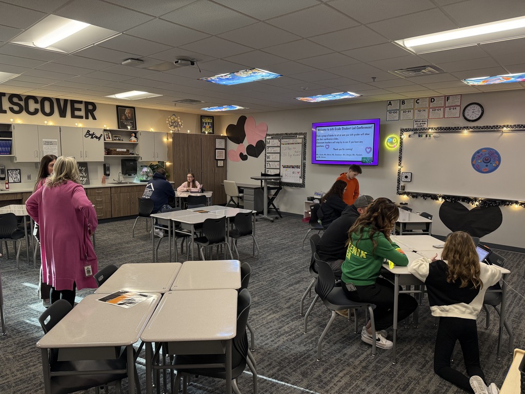 Students and parents sit at desks in a classroom chatting with teachers. 